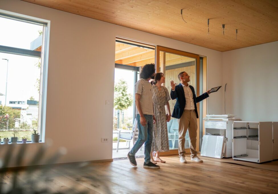 A professional Hispanic male real estate agent engages with a couple, presenting features of a spacious, sunlit home, while the diverse pair attentively follows along, considering potential ownership.